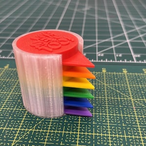 Puede incluir: Un objeto cilíndrico impreso en 3D con una carcasa exterior blanca translúcida y una pila de seis insertos triangulares en rojo, naranja, amarillo, verde, azul y morado. El inserto rojo superior presenta el texto en relieve "LIFT TO SORT".
