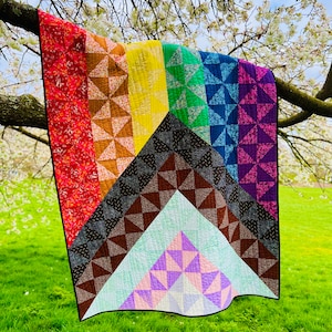 May include: A rainbow quilt with a geometric pattern of stars and triangles. The quilt is hanging from a tree branch in a grassy field.