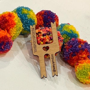 May include: A wooden pom pom maker with the words "WePomPom!" and "@weyarmtoo" engraved on it. The maker is surrounded by colourful pom poms.