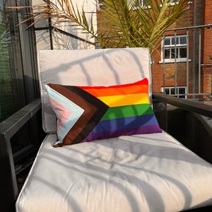 Op de afbeelding: Een wit kussen met een regenboog-pride-vlagkussen erop. Het kussen is gemaakt van stof met een patchwork-ontwerp. Het kussen ligt op een zwarte stoel op een balkon.
