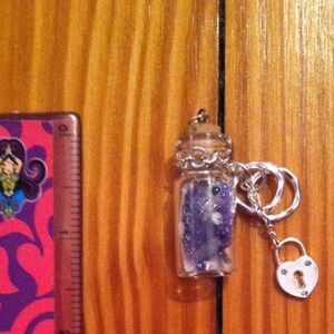 May include: A small glass bottle pendant filled with purple and white stones, topped with a cork and silver embellishments. Attached is a silver chain with a heart-shaped lock charm. A ruler with a colorful design is to the left.