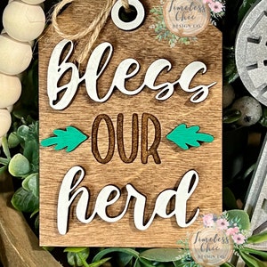 May include: Wooden sign with the text "Bless Our Herd" in white lettering. The sign has a rustic brown background with green leaves on either side of the word "our". The sign has a hole at the top for hanging.