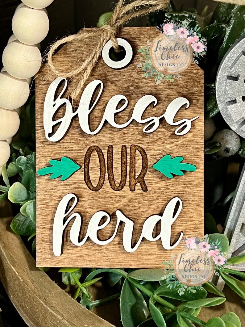 May include: Wooden sign with the text "Bless Our Herd" in white lettering. The sign has a rustic brown background with green leaves on either side of the word "our". The sign has a hole at the top for hanging.