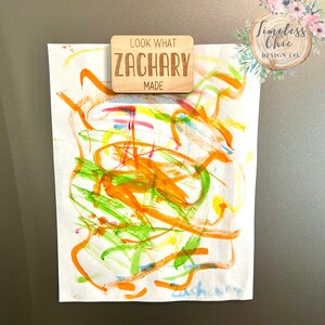 May include: A wooden sign with the text "Look What Zachary Made" on a white background. The sign is attached to a white sheet of paper with colorful abstract artwork on it.