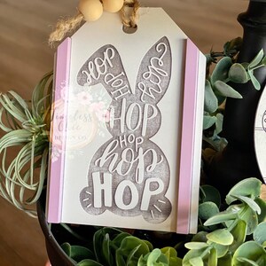 May include: A white wooden Easter bunny tag with the words "Hop Hop Hop Hop" written in gray. The tag has a pink ribbon on each side and is decorated with wooden beads.