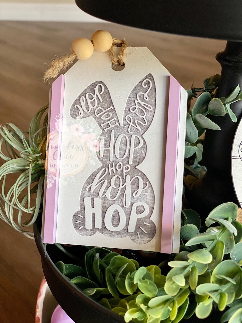 May include: A white wooden Easter bunny tag with the words "Hop Hop Hop Hop" written in gray. The tag has a pink ribbon on each side and is decorated with wooden beads.