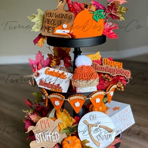 Pode incluir: Uma bandeja de tr&ecirc;s n&iacute;veis preta decorada com placas de madeira com tema de outono, um chap&eacute;u de tric&ocirc; e folhas de outono. As placas incluem as frases "You Want a Piece of Me?", "Gobble Til You Wobble", "I'm Stuffed", "Oh Snap!" e "I Love Fall Most of All".