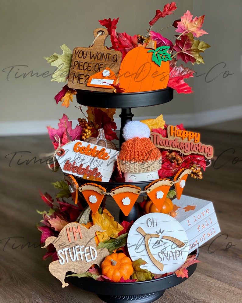 Pode incluir: Uma bandeja de tr&ecirc;s n&iacute;veis preta decorada com placas de madeira com tema de outono, um chap&eacute;u de tric&ocirc; e folhas de outono. As placas incluem as frases "You Want a Piece of Me?", "Gobble Til You Wobble", "I'm Stuffed", "Oh Snap!" e "I Love Fall Most of All".