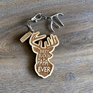Peut inclure: Porte-cl&eacute;s en bois avec une silhouette de cerf et le texte "Best Buckin' Papa Ever". Une petite &eacute;tiquette avec le nom "Zachary" est attach&eacute;e au porte-cl&eacute;s.