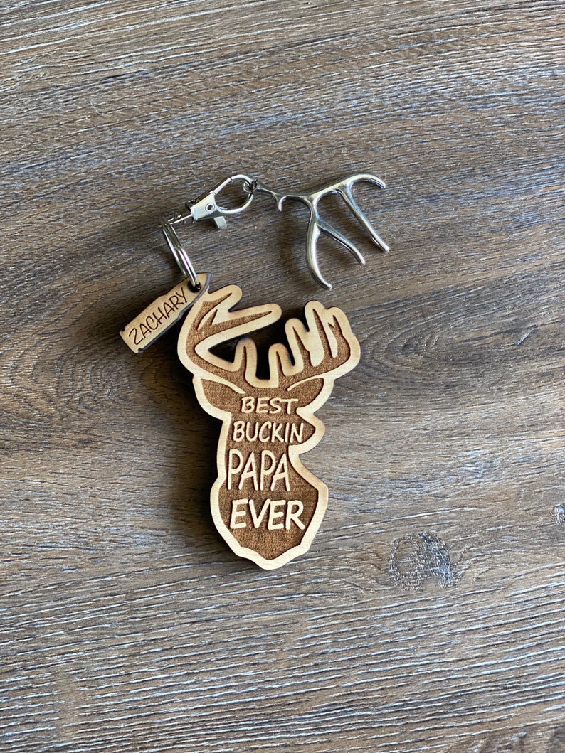 Peut inclure: Porte-cl&eacute;s en bois avec une silhouette de cerf et le texte "Best Buckin' Papa Ever". Une petite &eacute;tiquette avec le nom "Zachary" est attach&eacute;e au porte-cl&eacute;s.