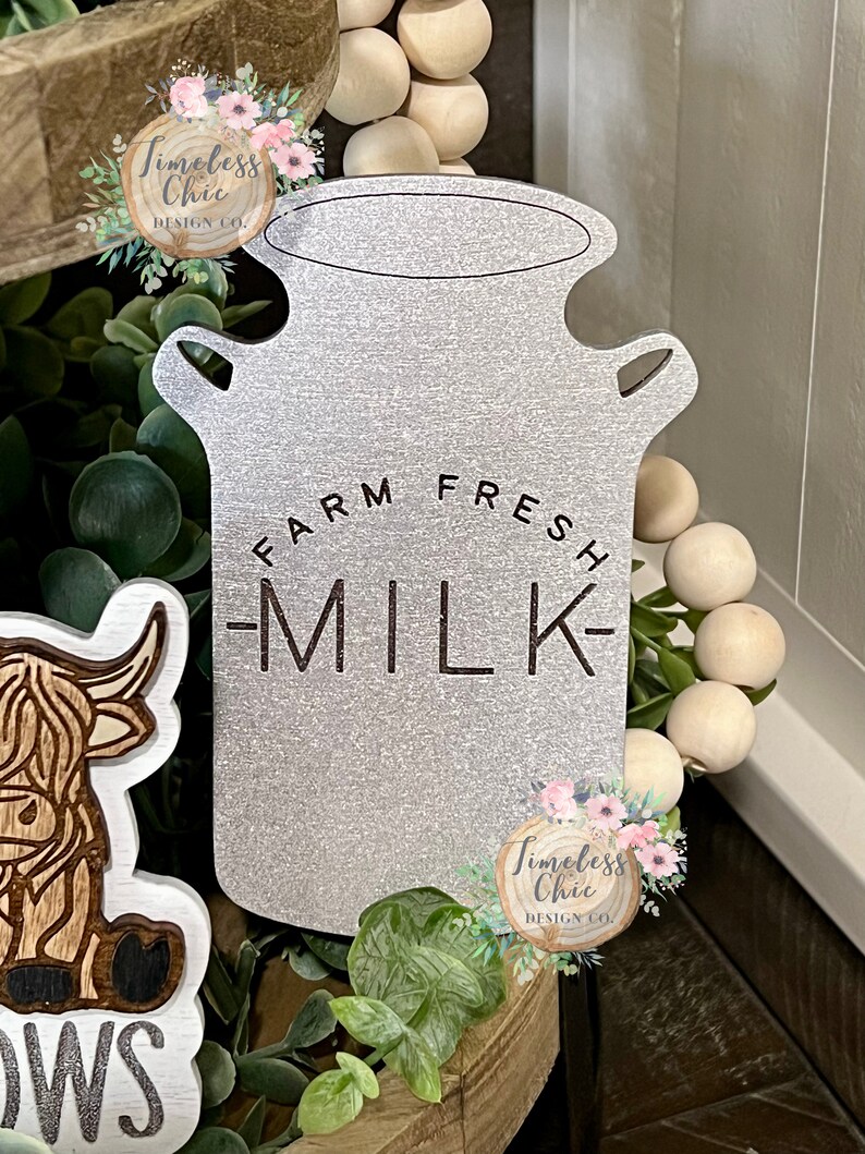 May include: A silver milk jug with the words "Farm Fresh Milk" on it. The jug is sitting on a tiered wooden tray with greenery and wooden beads.