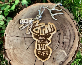 Könnte beinhalten: Holzschlüsselanhänger mit einer Hirsch-Silhouette und dem Text "Best Buckin' Dad Ever". Drei kleinere Holzanhänger mit den Namen "Zachary", "Mackenzie" und "Madison" sind am Schlüsselanhänger befestigt.