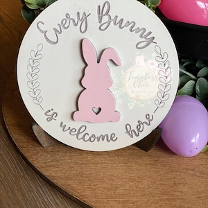 May include: A white wooden sign with the text "Every Bunny is welcome here" in gray script. A pink bunny silhouette with a heart is in the center of the sign.
