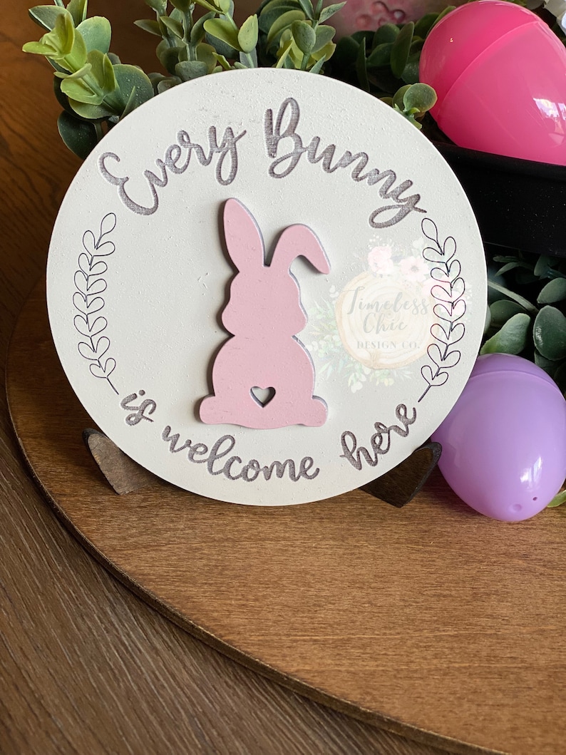 May include: A white wooden sign with the text "Every Bunny is welcome here" in gray script. A pink bunny silhouette with a heart is in the center of the sign.
