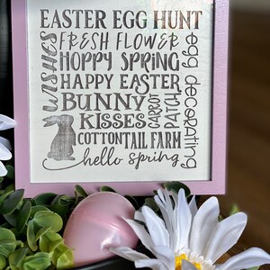 May include: A pink wooden sign with the text "EASTER EGG HUNT", "FRESH FLOWER", "HOPPY SPRING", "HAPPY EASTER", "BUNNY", "KISSES", "COTTONTAIL FARM", and "hello spring" in black lettering. The sign is decorated with a bunny silhouette.