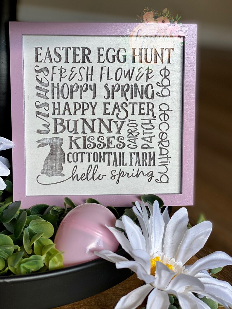May include: A pink wooden sign with the text "EASTER EGG HUNT", "FRESH FLOWER", "HOPPY SPRING", "HAPPY EASTER", "BUNNY", "KISSES", "COTTONTAIL FARM", and "hello spring" in black lettering. The sign is decorated with a bunny silhouette.