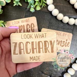 May include: Two wooden signs with the text "Look What Zachary Made" and "Look What [illegible] Made" in a wood-burned font. The signs are on a wooden surface with white beads in the background.