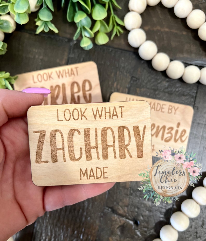 May include: Two wooden signs with the text "Look What Zachary Made" and "Look What [illegible] Made" in a wood-burned font. The signs are on a wooden surface with white beads in the background.