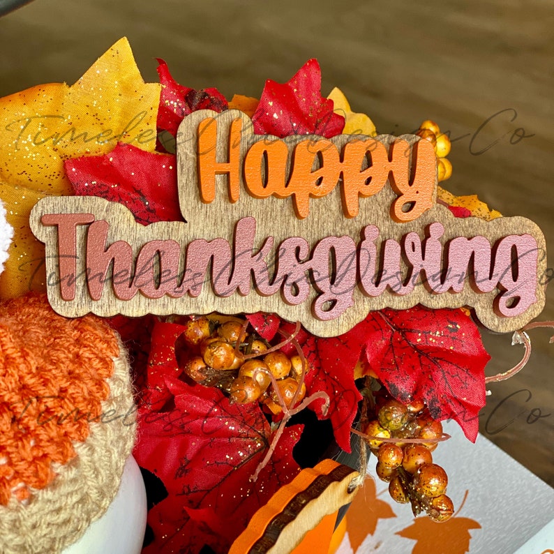 Pode incluir: Uma placa de madeira com as palavras "Happy Thanksgiving" em letras laranja e rosa. A placa &eacute; cercada por folhagem de outono, incluindo folhas vermelhas e amarelas e bagas marrons.