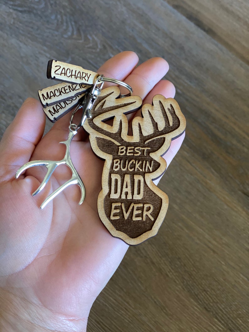 Peut inclure: Porte-cl&eacute;s en bois avec un motif de t&ecirc;te de cerf et le texte "Best Buckin' Dad Ever". Le porte-cl&eacute;s comprend &eacute;galement trois petites &eacute;tiquettes en bois avec les noms "Zachary", "Mackenzie" et "Madison".