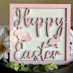 May include: A pink wooden sign with the words "Happy Easter" in white lettering. The sign is decorated with two pink bunny rabbits.