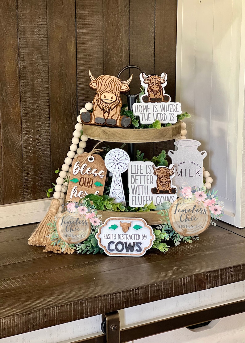 May include: A tiered tray decorated with wooden signs featuring cow and farm-themed sayings, including "Home is where the herd is", "Life is better with fluffy cows", and "Easily distracted by cows". The tray is also decorated with a wooden windmill, a milk jug, and a wooden bead garland.