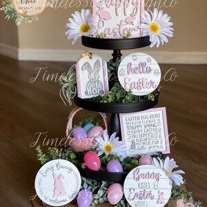 May include: A tiered tray decorated for Easter with pink and white signs, flowers, and eggs. The signs say "Happy Easter", "Hello Easter", "Every Bunny is Welcome Here", and "Bunny Kisses & Easter Wishes".