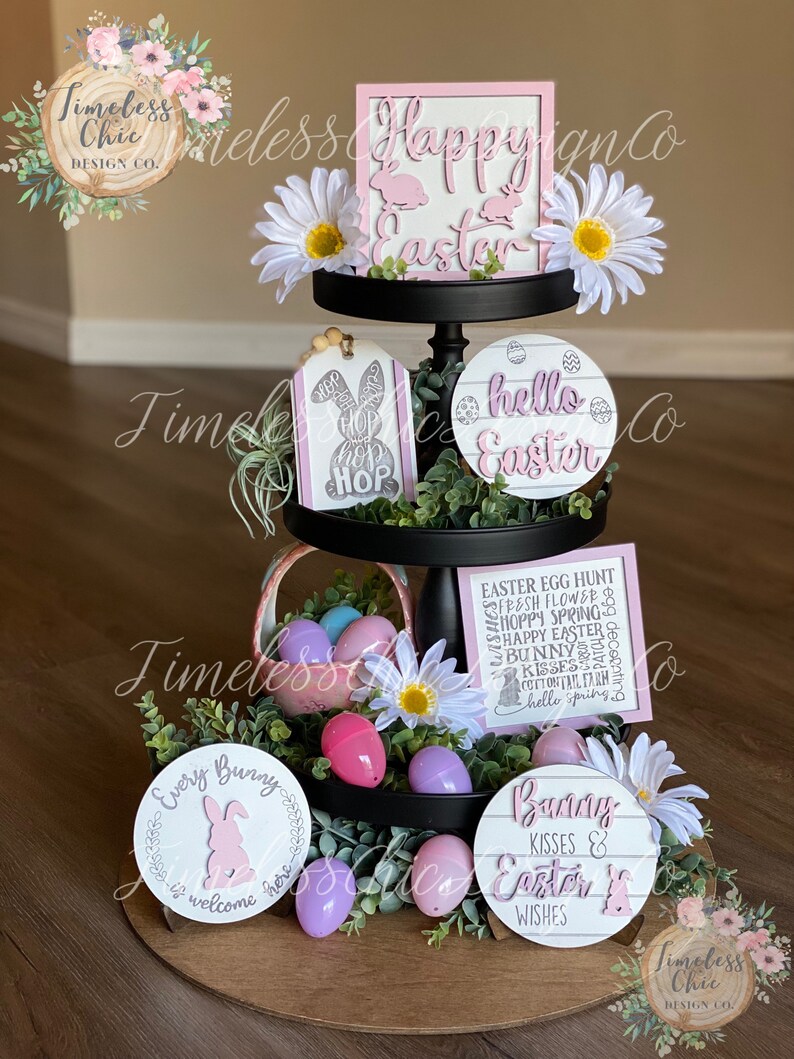 May include: A tiered tray decorated for Easter with pink and white signs, flowers, and eggs. The signs say "Happy Easter", "Hello Easter", "Every Bunny is Welcome Here", and "Bunny Kisses & Easter Wishes".