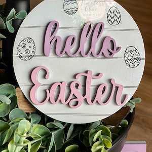 May include: A white wooden round sign with a pink "hello easter" message. The sign has a distressed wood look and is decorated with black and white patterned eggs.