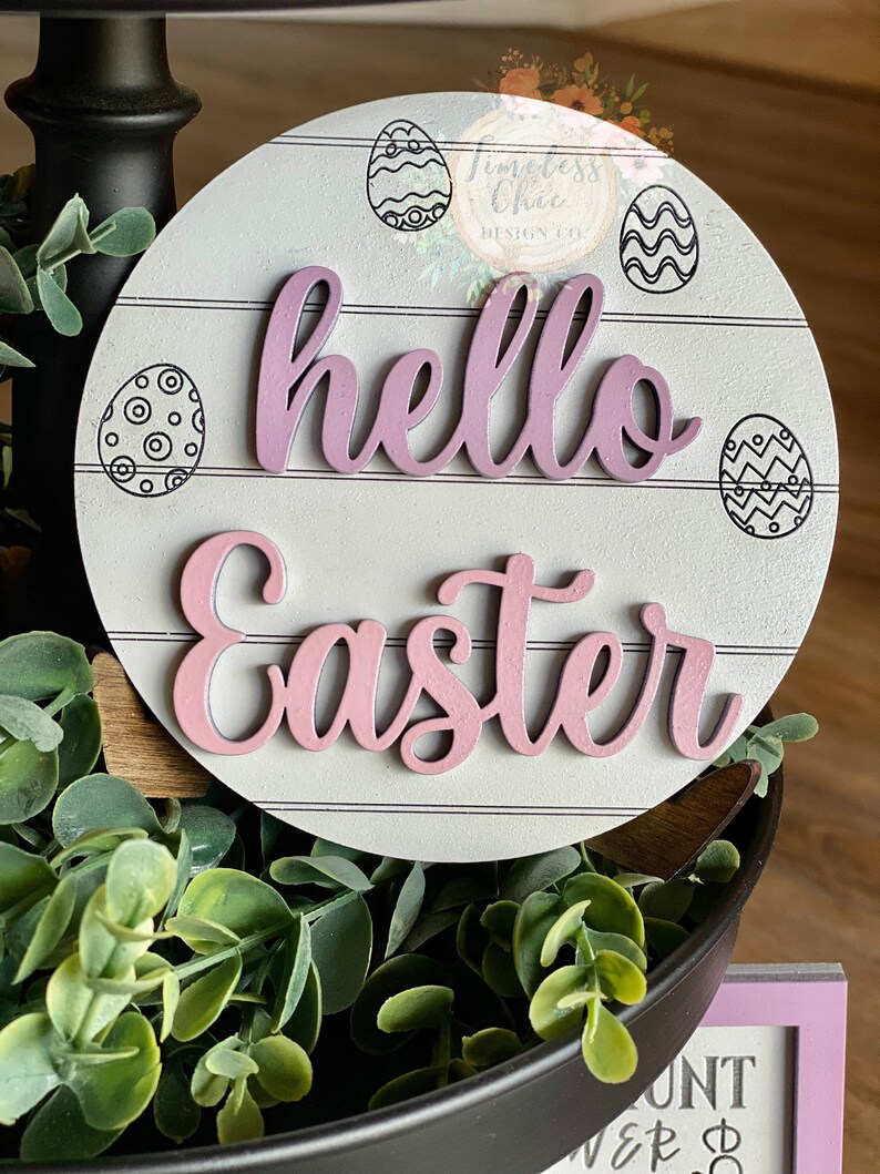 May include: A white wooden round sign with a pink "hello easter" message. The sign has a distressed wood look and is decorated with black and white patterned eggs.