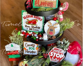 May include: A three-tiered tray decorated for Christmas with various signs, including "Santa's Milk & Cookies", "Sleigh Rides", "Carrots for Reindeer", "Santa Stop Here", and "Mail for Santa Claus The North Pole". The tray is decorated with greenery, red berries, and a small gnome.