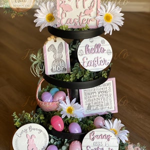 May include: A three-tiered tiered tray decorated for Easter. The top tier features a pink and white sign that says "Happy Easter". The middle tier has a pink and white tag that says "Hop Hop Hop" and a white sign with the words "hello Easter". The bottom tier has two round signs, one that says "Every Bunny is welcome here" and the other that says "Bunny Kisses & Easter Wishes". The tray is decorated with white flowers, greenery, and pastel-colored plastic eggs.