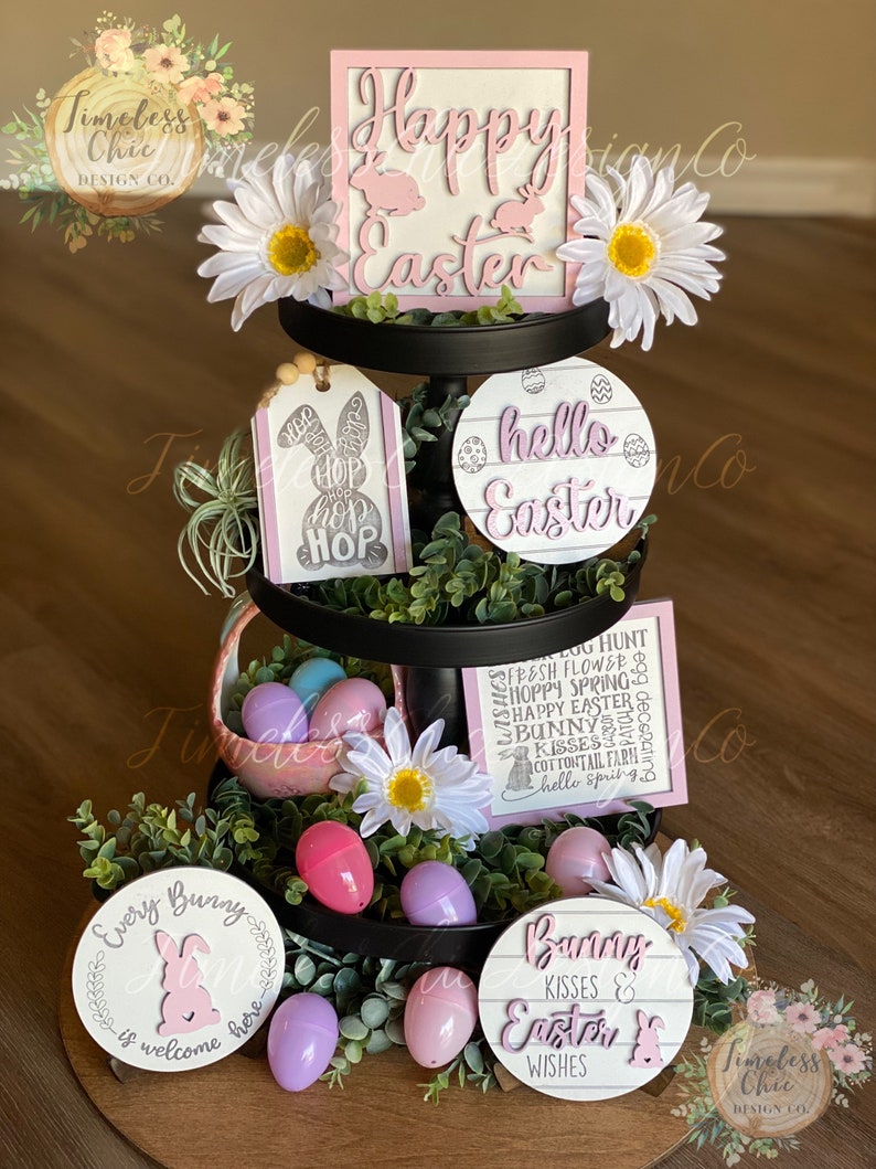 May include: A three-tiered tiered tray decorated for Easter. The top tier features a pink and white sign that says "Happy Easter". The middle tier has a pink and white tag that says "Hop Hop Hop" and a white sign with the words "hello Easter". The bottom tier has two round signs, one that says "Every Bunny is welcome here" and the other that says "Bunny Kisses & Easter Wishes". The tray is decorated with white flowers, greenery, and pastel-colored plastic eggs.
