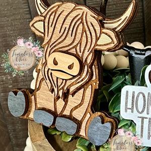 May include: A wooden cutout of a Highland cow with a brown and white striped pattern. The cow is sitting with its legs tucked under its body. The cutout is painted with a light blue color on the hooves.