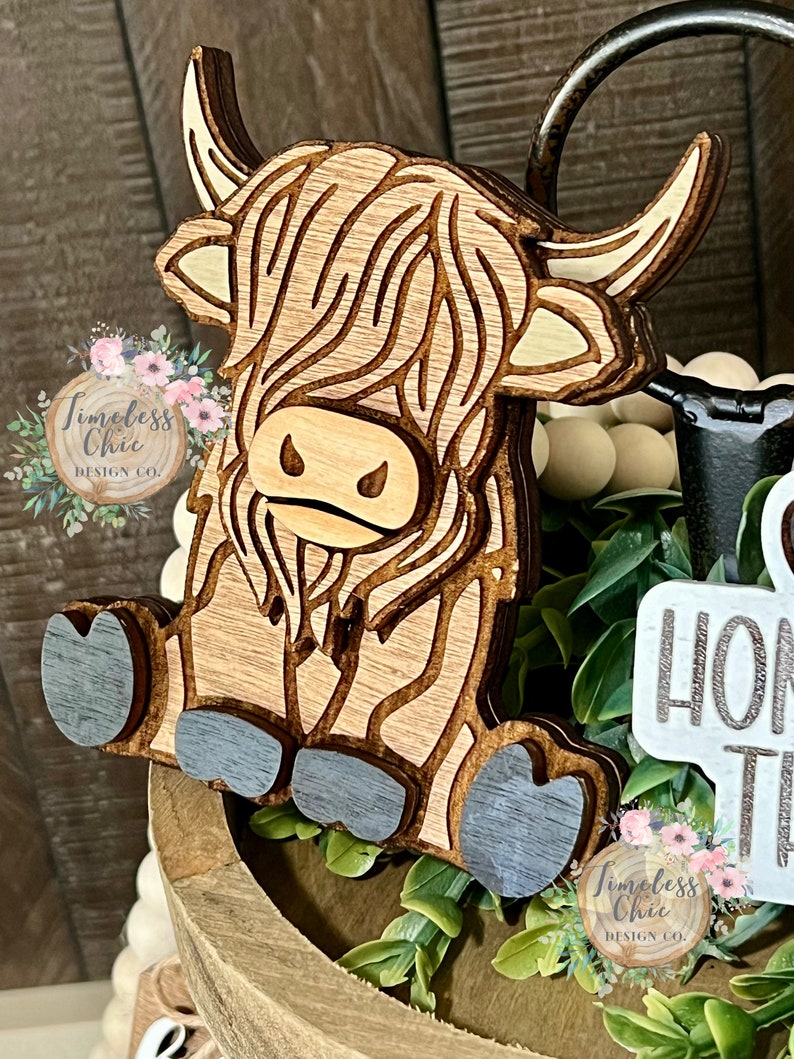May include: A wooden cutout of a Highland cow with a brown and white striped pattern. The cow is sitting with its legs tucked under its body. The cutout is painted with a light blue color on the hooves.