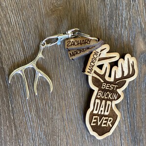 Peut inclure: Porte-cl&eacute;s en bois avec une silhouette de cerf et le texte "Best Buckin' Dad Ever". Trois &eacute;tiquettes en bois avec les noms "Zachary", "Mackenzie" et "Madison" sont attach&eacute;es au porte-cl&eacute;s.