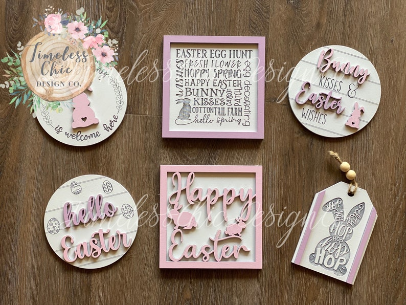 May include: A set of six Easter-themed wooden signs with pink and white paint. The signs feature various Easter-related phrases and images, including bunnies, eggs, and the words "Happy Easter", "Bunny Kisses & Easter Wishes", and "Hello Easter".