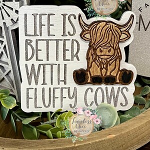 May include: Wooden sign with the text "Life is better with fluffy cows" in silver lettering. A brown and white highland cow is depicted on the sign.