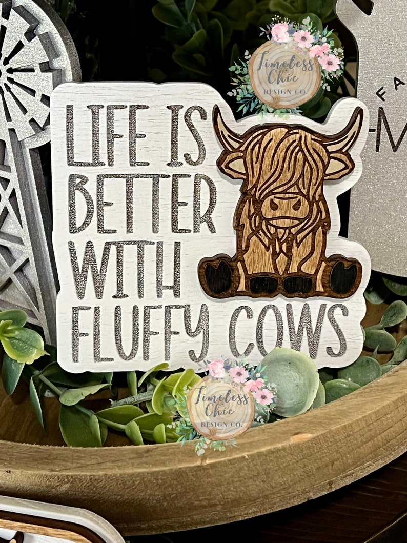 May include: Wooden sign with the text "Life is better with fluffy cows" in silver lettering. A brown and white highland cow is depicted on the sign.