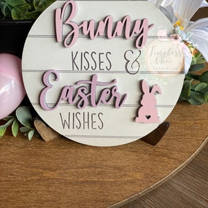 May include: A pink and white wooden sign with the text "Bunny Kisses & Easter Wishes" and a pink bunny rabbit with a heart on its chest. The sign is on a wooden surface with green plants and white flowers.