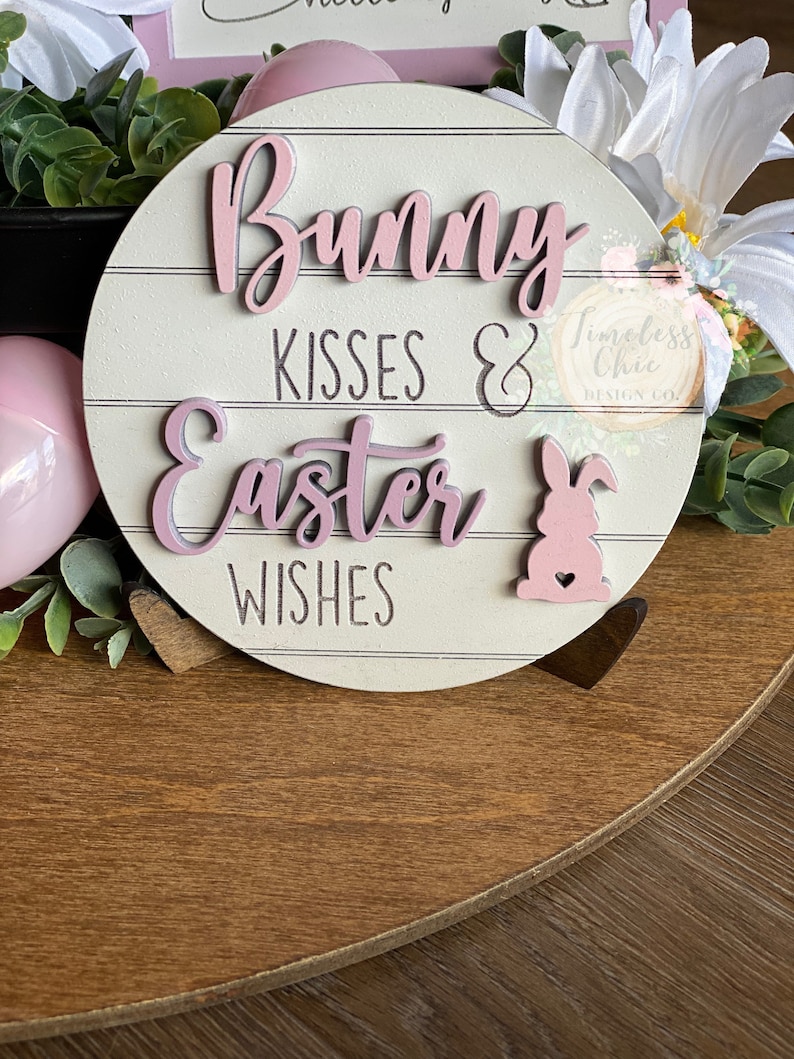May include: A pink and white wooden sign with the text "Bunny Kisses & Easter Wishes" and a pink bunny rabbit with a heart on its chest. The sign is on a wooden surface with green plants and white flowers.