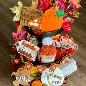 Pode incluir: Uma bandeja de tr&ecirc;s n&iacute;veis decorada com placas de madeira com tema de outono, incluindo "Voc&ecirc; quer um peda&ccedil;o de mim?", "Gobble 'Til You Wobble", "Estou cheio", "Feliz Dia de A&ccedil;&atilde;o de Gra&ccedil;as", "Eu amo o outono mais do que tudo", e "Oh Snap!" A bandeja &eacute; decorada com folhas de outono, ab&oacute;boras e um gnomo de croch&ecirc;.