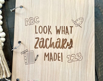 May include: A wooden scrapbook with a light brown wood grain. The cover has the text "ABC LOOK WHAT Zachary MADE! 123" with a drawing of a paintbrush and palette and a pencil and notebook. The scrapbook has silver rings.