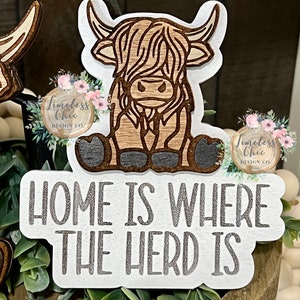 May include: A white wooden sign with the text "Home is where the herd is" in silver glitter lettering. A brown and white cartoon Highland cow is sitting on the sign.