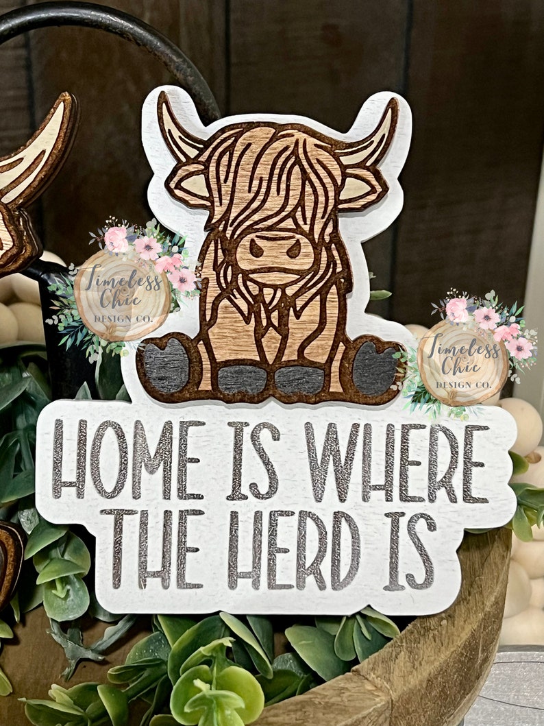 May include: A white wooden sign with the text "Home is where the herd is" in silver glitter lettering. A brown and white cartoon Highland cow is sitting on the sign.