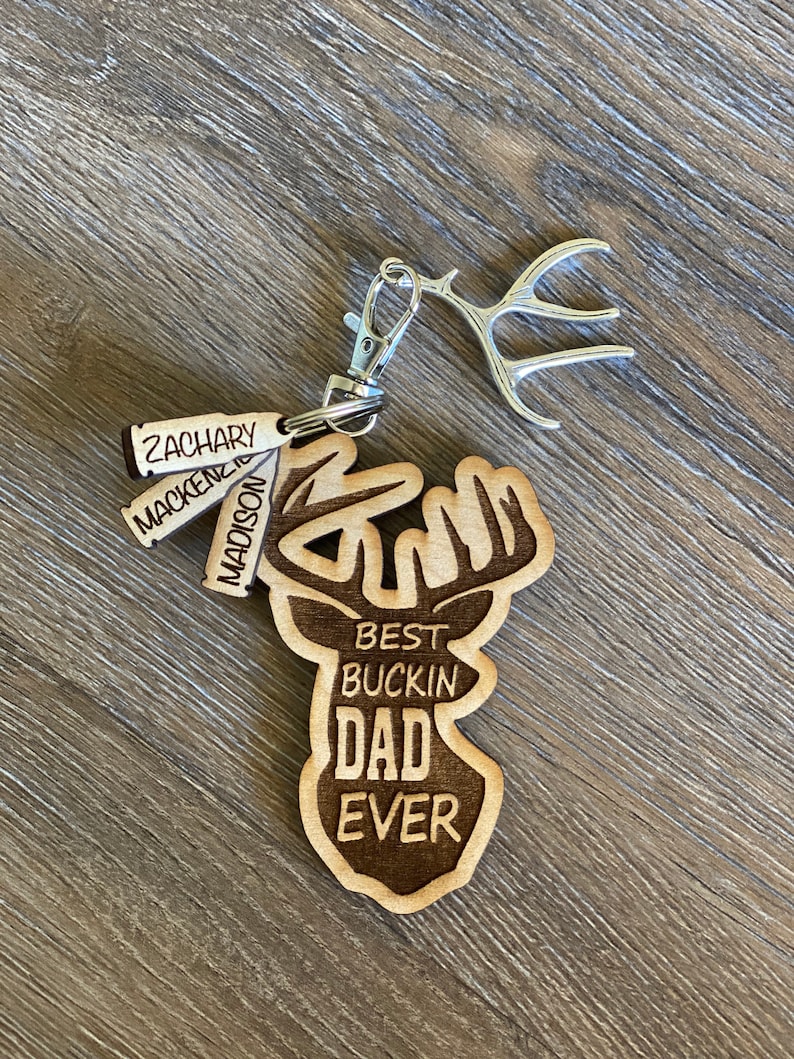 Peut inclure: Porte-cl&eacute;s en bois avec un motif de t&ecirc;te de cerf. La t&ecirc;te de cerf est grav&eacute;e du texte "Best Buckin' Dad Ever". Le porte-cl&eacute;s comprend &eacute;galement quatre petites &eacute;tiquettes en bois grav&eacute;es avec les noms "Zachary", "Mackenzie", "Maddox" et "Nason".