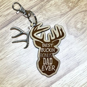 Peut inclure: Porte-cl&eacute;s en bois en forme de t&ecirc;te de cerf avec des bois. Le porte-cl&eacute;s porte l'inscription "Best Buckin' Bonus Dad Ever".