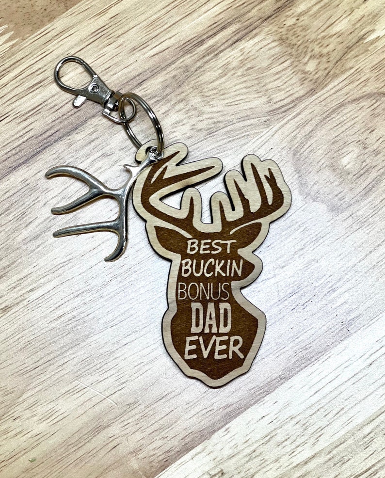 Peut inclure: Porte-cl&eacute;s en bois en forme de t&ecirc;te de cerf avec des bois. Le porte-cl&eacute;s porte l'inscription "Best Buckin' Bonus Dad Ever".