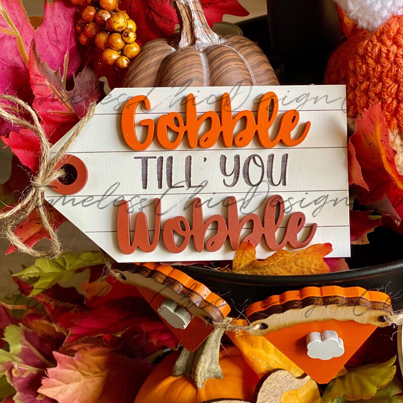 Pode incluir: Uma placa de madeira branca com o texto "Gobble Till' You Wobble" em letras laranja e pretas. A placa &eacute; decorada com folhas de outono e ab&oacute;boras.