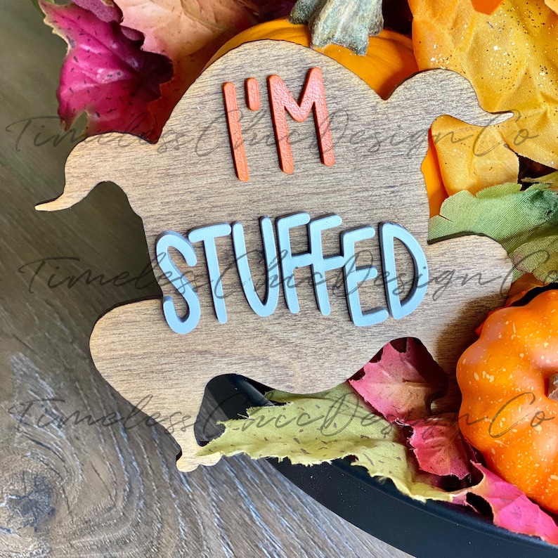 Pode incluir: Um recorte de peru de madeira com as palavras "I'm Stuffed" em letras azuis e laranja. O peru est&aacute; rodeado de folhas de outono e ab&oacute;boras.